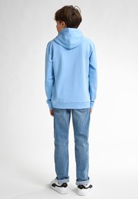 Hanorac de culoare albastru deschis cu glugă ajustabilă, din material din bumbac, cu manșete din tricot, purtat împreună cu blugi albaștri și adidași albi cu accente negre.
