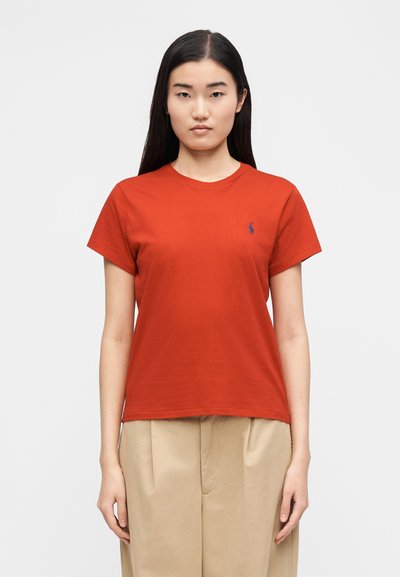 Piros pamutpóló kerek nyakkivágással és rövid ujjakkal, bal mellkasán kis kék logóval. Sima textúrájú, lezser szabású.