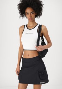 Fehér cropped top fekete szélekkel és logóval; sötét szoknya cipzárral és zsebekkel; modell egy kis fekete táskát tart.
