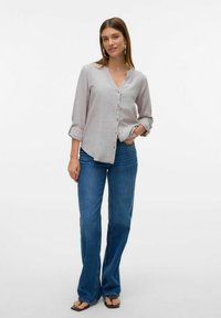 Chemise gris clair à col en V, avec des manches retroussées, associée à un jean bleu évasé. La tenue est complétée par des sandales plates noires.