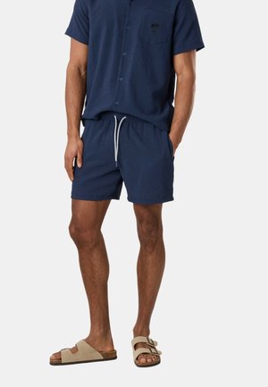 Homme portant une chemise à manches courtes texturée bleu marine avec des boutons et un short assorti avec un cordon de serrage blanc, sandales beige à double bride.