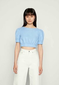 Mulher jovem com cabelo liso e escuro, vestindo um top cropped de mangas bufantes azul claro e calças brancas de cintura alta, de pé contra um fundo claro.