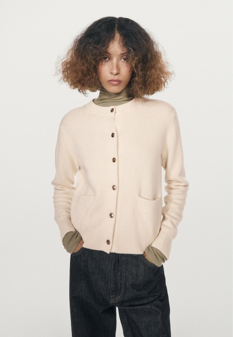 Beige knit cardigan with a round neckline, front button closure, two pockets, and ribbed cuffs, layered over a green top.