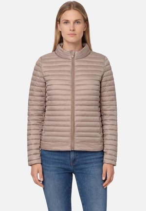 Donna con capelli biondi lisci che indossa una giacca trapuntata orizzontale color beige chiaro chiusa con la cerniera e jeans blu, in piedi contro uno sfondo semplice.