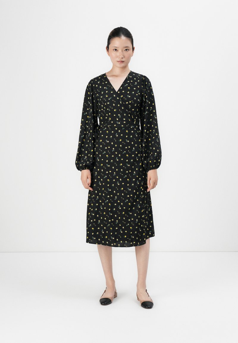 Black wrap dress with yellow floral print, long puff sleeves, and a mid-length hem. Smooth fabric texture, complemented by simple black flats.