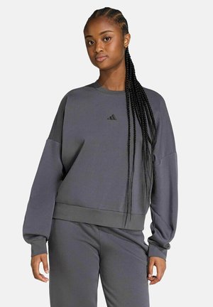 Frau mit langem geflochtenem Haar, die einen lockeren dunkelgrauen Adidas-Sweatshirt und passende Hose trägt und vor einem einfarbigen Hintergrund steht.