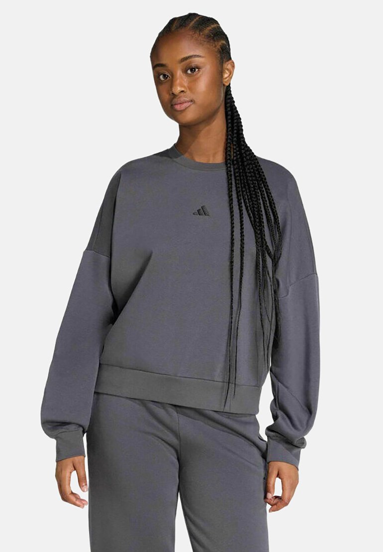Frau mit langem geflochtenem Haar, die einen lockeren dunkelgrauen Adidas-Sweatshirt und passende Hose trägt und vor einem einfarbigen Hintergrund steht.
