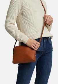 Bruine leren schoudertas met een gestructureerde afwerking, compacte rechthoekige vorm en een verstelbare schouderriem, gedragen door een persoon in denim.