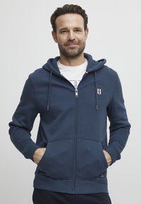 Hoodie zippé bleu marine avec une poche avant, des poignets côtelés et un patch logo sur la poitrine gauche. Tissu doux et capuche ajustable avec cordon.