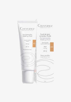 Korrigierende Make-up-Flüssigkeit in einer weißen Tube mit geschwungenen Designelementen. Verfügt über SPF 20, einen hellen Honigton und eine Verpackung mit mehrsprachigem Text.