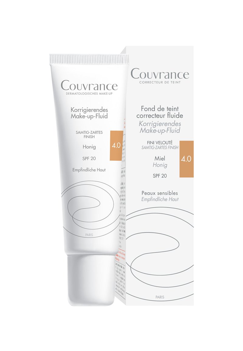 Korrigierende Make-up-Flüssigkeit in einer weißen Tube mit geschwungenen Designelementen. Verfügt über SPF 20, einen hellen Honigton und eine Verpackung mit mehrsprachigem Text.