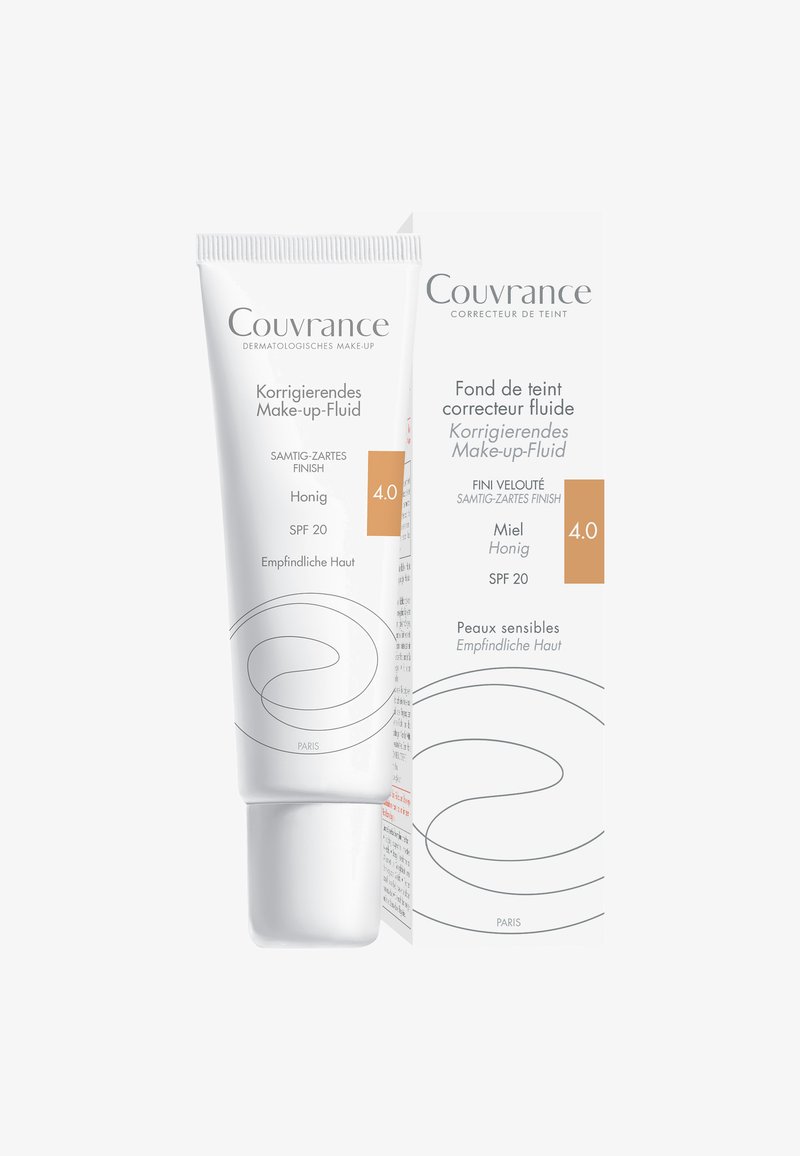 Korrigierende Make-up-Flüssigkeit in einer weißen Tube mit geschwungenen Designelementen. Verfügt über SPF 20, einen hellen Honigton und eine Verpackung mit mehrsprachigem Text.