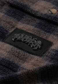 Tessuto flanella a quadri in toni terrosi di marrone e blu navy, con una toppa in pelle nera testurizzata con design impresso.