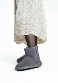 Stivali alla caviglia in suede grigio con punta rotonda, morbida fodera in pelliccia e una decorazione a bottone. Indossati con una gonna leggera a fantasia e collant neri.