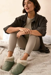 Grønne quiltede hjemmesko med pelskant, båret af en person i beige ribbede leggings og en mørk fløjlsk Jakke, siddende på et struktureret tæppe.