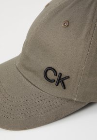 Khaki honkbalpet met een gestructureerd ontwerp, voorzien van een zwart geborduurd "CK"-logo, gestructureerde stof en gestikte ventilatiegaatjes.