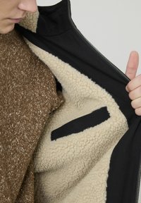 Veste en tissu brun avec une fermeture éclair noire et doublure beige en laine, dotée d'une poche cachée à l'intérieur.