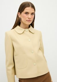 Beige cropped Jacke mit runder Kragen, fünf Knöpfen und einer Fronttasche. Hergestellt aus glattem Stoff mit einem strukturierten Design.