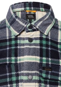 Nærbillede af en navy, hvid og grønternet fleece-skjorte med krave, sorte trykknapper og Camel Active mærkeetiket på lommen og indeni kraven.
