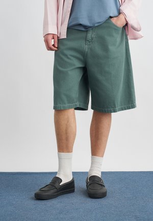 Άτομο που φοράει πράσινα σορτς μέχρι το γόνατο, λευκές κάλτσες κάλτσα, μαύρα παπούτσια slip-on, μπλε πουκάμισο και ανοιχτό ροζ πουκάμισο με κουμπιά.