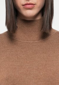 Brun genser med høy hals laget av ribbestrikket materiale. Nærbilde viser tekstur og design på halsen, og fremhever dens tettsittende form.