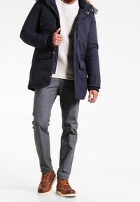 Marinblå vadderad jacka med huva, krämfärgad stickad tröja, grå skräddarsydda byxor och bruna snörkängor med vita sulor.