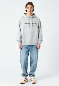 Felpa grigia con cappuccio e testo nero, tasca a marsupio frontale, abbinata a jeans azzurri chiari e sneakers beige con accenti rossi e blu navy.