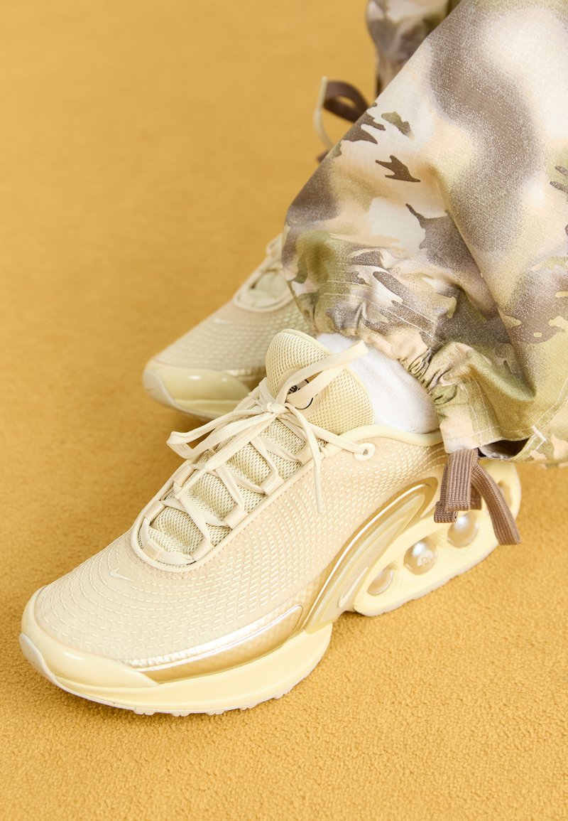 Beige sportliche Sneakers mit einem strukturierten Mesh-Obermaterial, runder Zehenform und sichtbaren Luftpolstern. Kombiniert mit gemusterten Jogginghosen.
