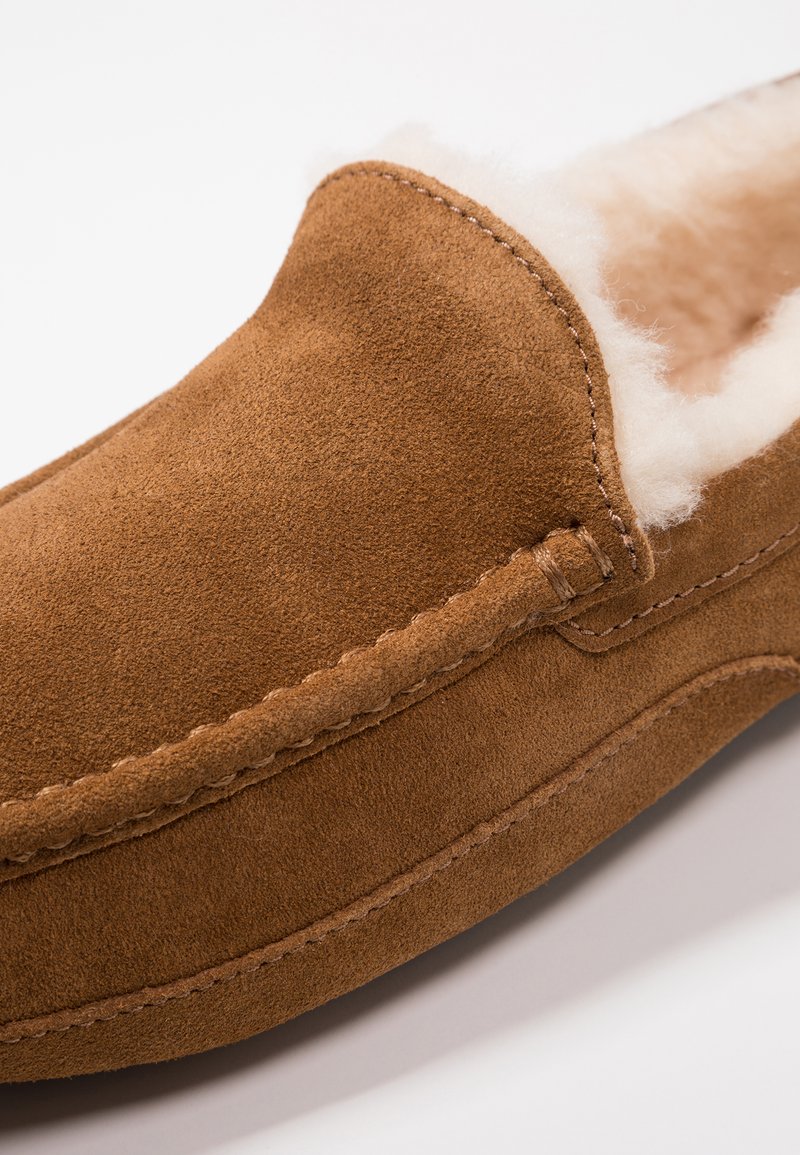 Brown suede slipper with a fluffy cream lining, rounded toe, and subtle stitching details along the edges for added texture.