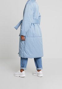 Cappotto imbottito blu chiaro con vita cintrata, texture trapuntata, maniche lunghe e design di lunghezza media, abbinato a jeans in denim arrotolati e sneaker bianche.