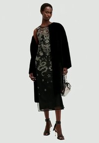 Fekete estélyi ruha, amelyet összetett ezüst hímzések borítanak kígyókkal, csillagokkal és pillangókkal; egy fekete műszőrme kabáttal és pántos magassarkú cipővel párosítva.