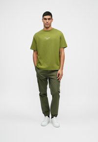 Olijfgroene katoenen t-shirt met korte mouwen en logo, gecombineerd met olijfgroene cargo broek met elastische boorden en witte sneakers.