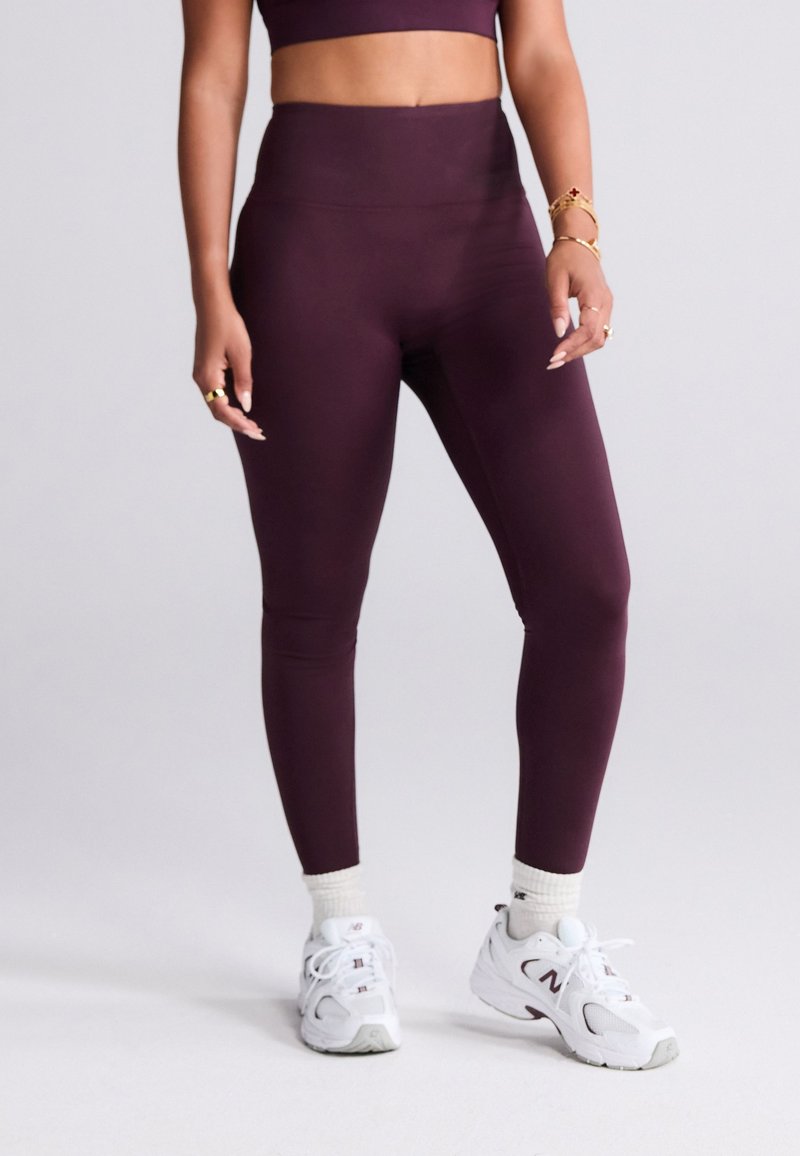 Dunkelrote Leggings mit hoher Taille aus dehnbarem Stoff, mit glatter Textur. Kombiniert mit weißen Turnschuhen und cremefarbenen Socken.