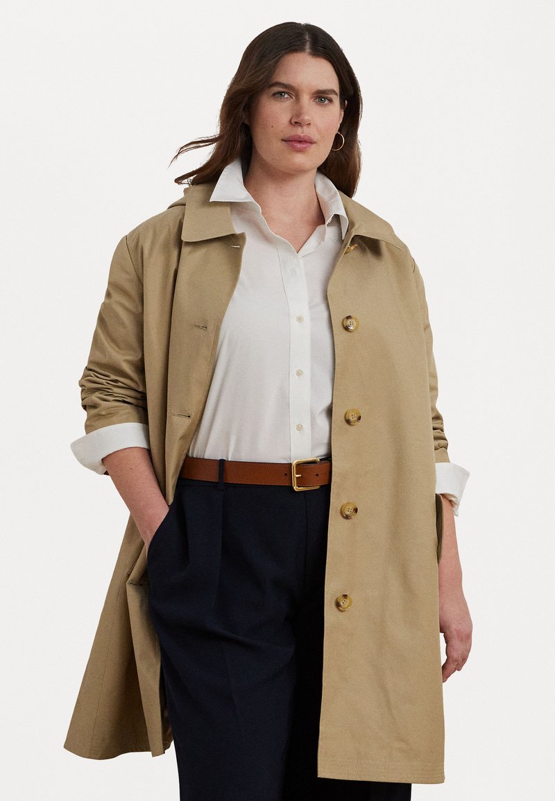 Beige trenchcoat med krage och knappstängning, i kombination med en vit skjorta, brun bälte och marinblå byxor. Slät tyg och skräddarsydd passform.
