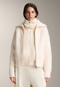 Fleecová bunda v krémovej farbe s vysokým golierom a zapínaním na zips. Textúrovaný vonkajší materiál a rebrované manžety. Nosená cez priliehavý krémový rolák.