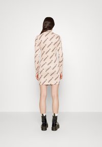 Lange mouwen, nauwsluitende jurk in lichtbeige met een herhaald bruin Adidas-logo patroon. Het model draagt zwarte enkellaarzen met een dikke zool.