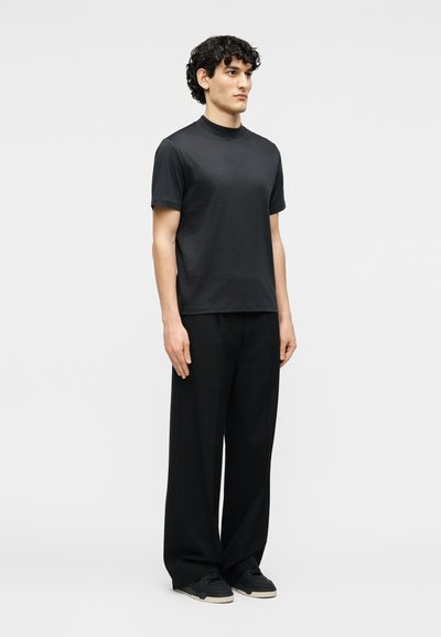 Νέος άντρας που στέκεται, φοράει απλό μαύρο κοντομάνικο πουκάμισο, φαρδιά μαύρα παντελόνια και μαύρα αθλητικά παπούτσια σε απλό λευκό φόντο.