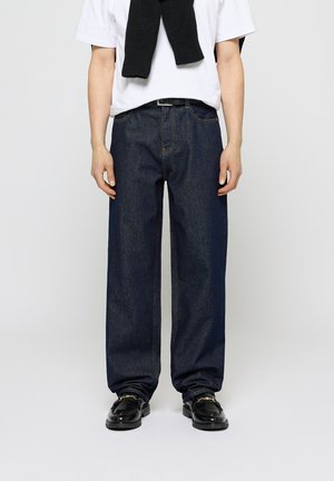 Osoba ubrana w luźne ciemnoniebieskie dżinsy, białą koszulkę, czarny sweter zarzucony na ramiona oraz czarne skórzane mokasyny z detalem w postaci złotego łańcuszka.