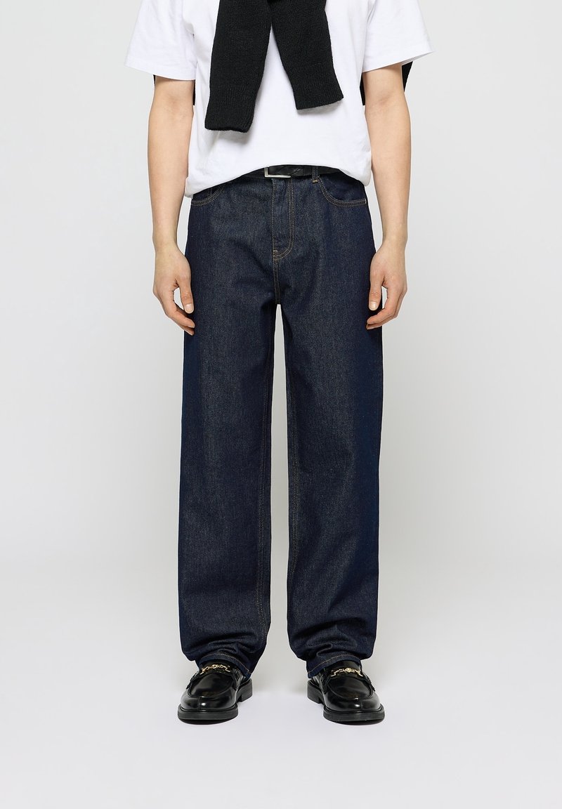 Personne portant un jean bleu foncé ample, un T-shirt blanc, un pull noir drapé sur les épaules, et des mocassins en cuir noirs avec un détail de chaîne dorée.