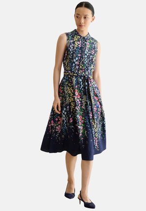 Frau trägt ein ärmelloses marineblaues Blumenkleid mit Kragen und Gürtel, kombiniert mit marineblauen Slingback-Absätzen, steht vor weißem Hintergrund.