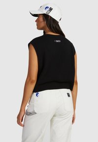 Black knit sleeveless top with ribbed hem, worn with white pants featuring patch pockets and contrasting blue and black accents. White cap displayed.