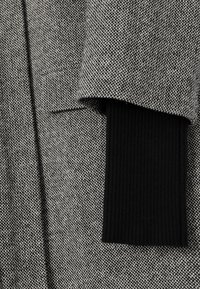 Tessuto in misto lana grigio e nero con un motivo testurizzato. Presenta un risvolto in maglia a coste nera all'apertura delle maniche.