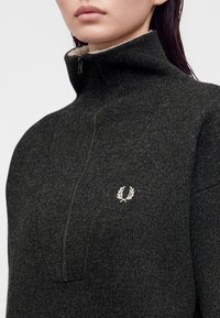 Gros plan d'une personne portant un pull en maille gris foncé à fermeture éclair avec un col haut et un petit logo blanc de couronne de laurier sur la poitrine.