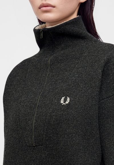 Gros plan d'une personne portant un pull en maille gris foncé à fermeture éclair avec un col haut et un petit logo blanc de couronne de laurier sur la poitrine.