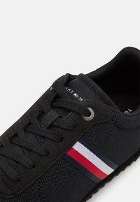 Czarne sneakersy z czarnymi sznurowadłami, tekstura materiału oraz czerwono-biało-granatowy pasek po boku w pobliżu sznurowadeł.