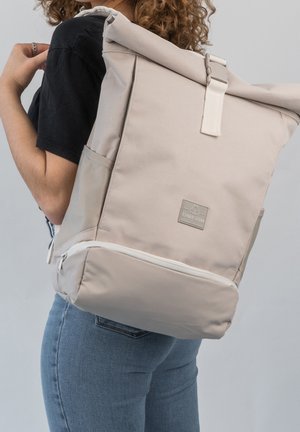 Personne portant un jean bleu et une chemise noire, portant un grand sac à dos beige à rabat roulé avec une poche latérale et un compartiment zippé en bas.