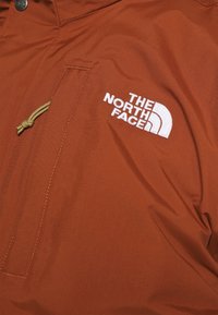 Hnědá anoraková bunda s jemnou texturou, vybavená kapsou na zip a vyšívaným bílým logem. Stahovací šňůrka v oblasti krku.