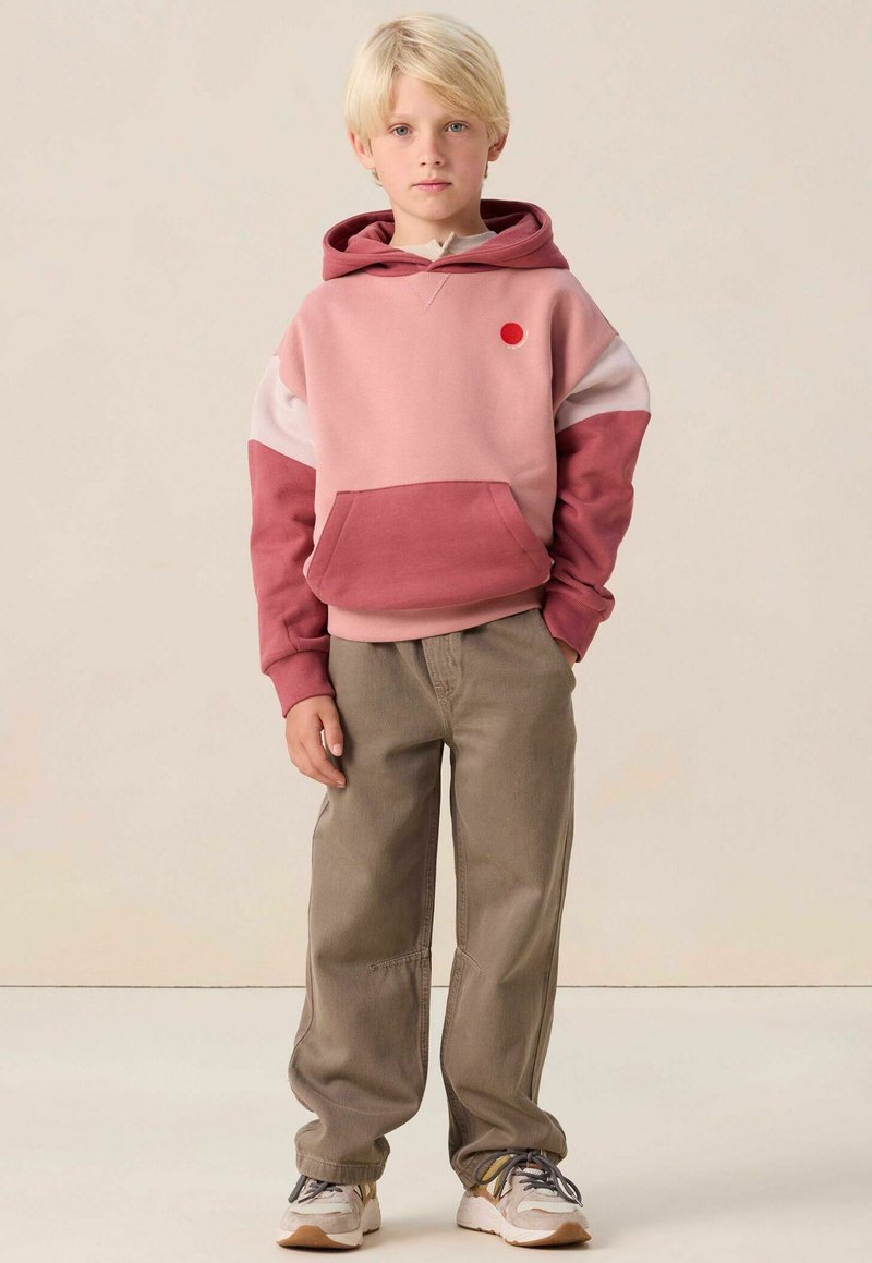 Sweat-shirt à capuche rose et marron avec une poche avant et un point d'accent rouge ; associé à un pantalon beige ample et des baskets grises.