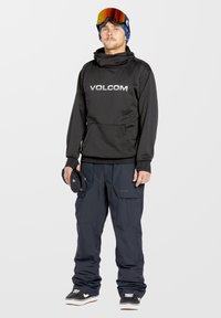 Sudadera con capucha negra con el logo de "VOLCOM", combinada con pantalones de nieve holgados de color azul oscuro y zapatillas negras. Lleva gafas de esquí en la frente.