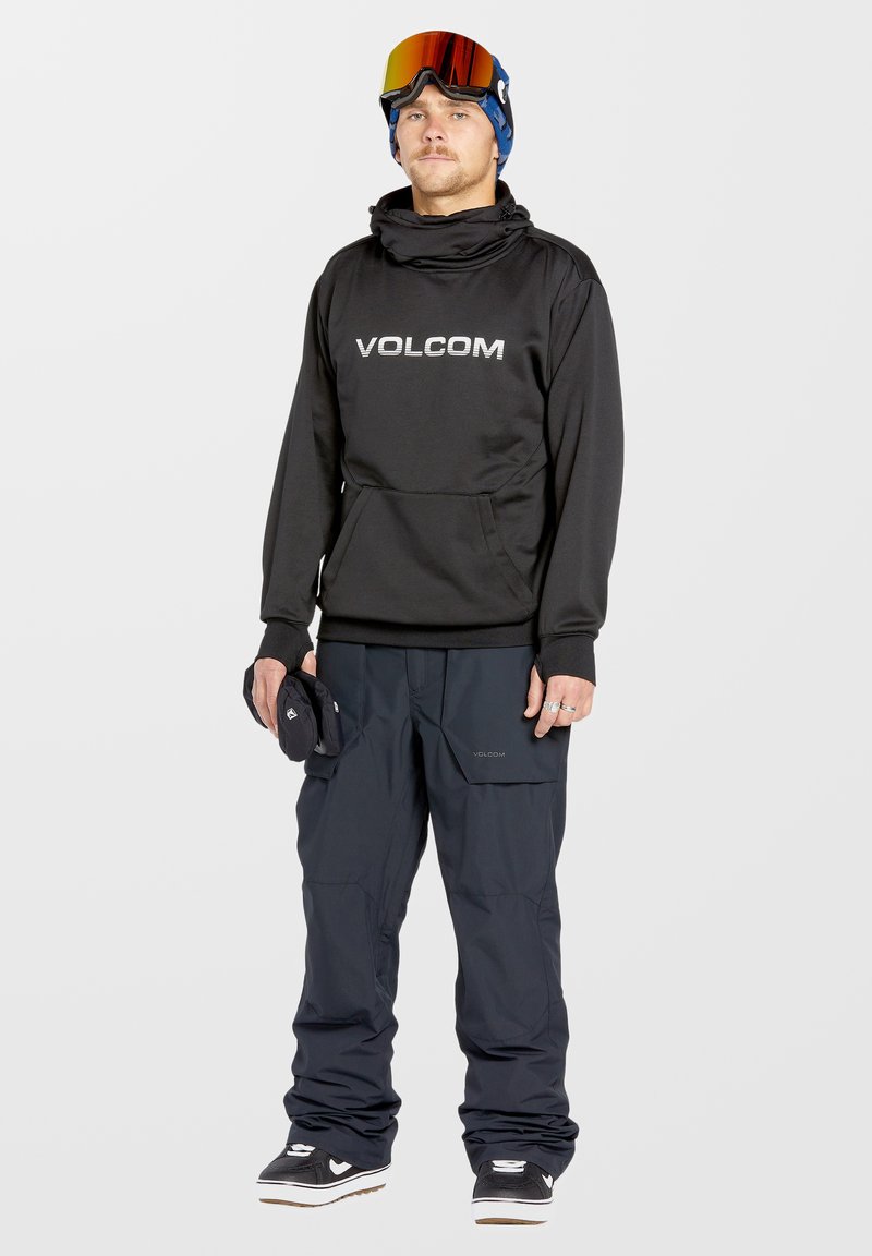 Sudadera con capucha negra con el logo de "VOLCOM", combinada con pantalones de nieve holgados de color azul oscuro y zapatillas negras. Lleva gafas de esquí en la frente.